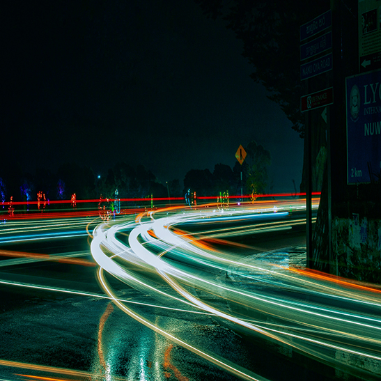 Bei Nacht fahren Autos über eine Straße. Ihre Lichter hinterlassen bunte Spuren: weiß, grün und rot. Die Straße ist nass, und im Hintergrund stehen Bäume und Schilder.